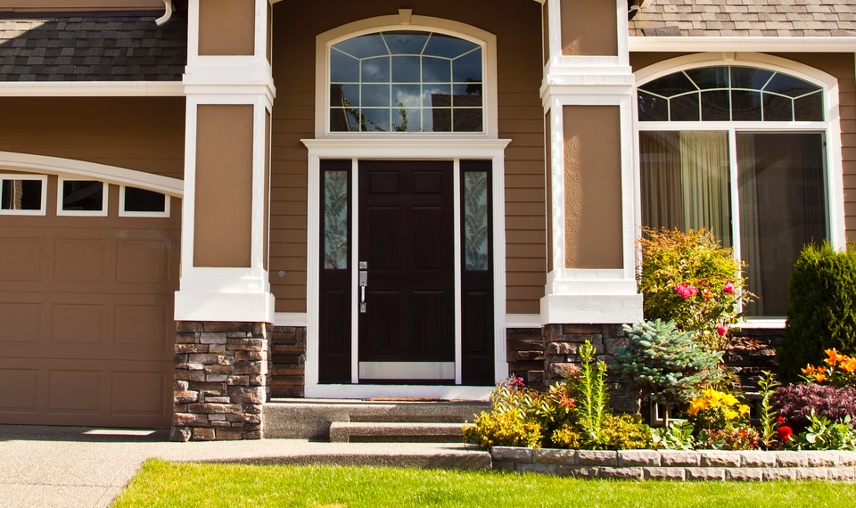 Porte exterieure moderne en bois foncé avec fenêtres latérales, encadrée par des colonnes blanches et une façade beige, située à l’entrée d’une maison avec jardin paysager.