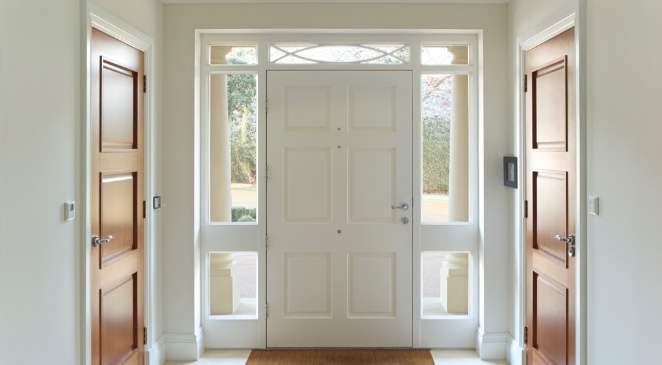 Entrée avec porte exterieure blanche, encadrée par des fenêtres latérales, offrant une luminosité naturelle, et deux portes intérieures en bois placées de chaque côté.